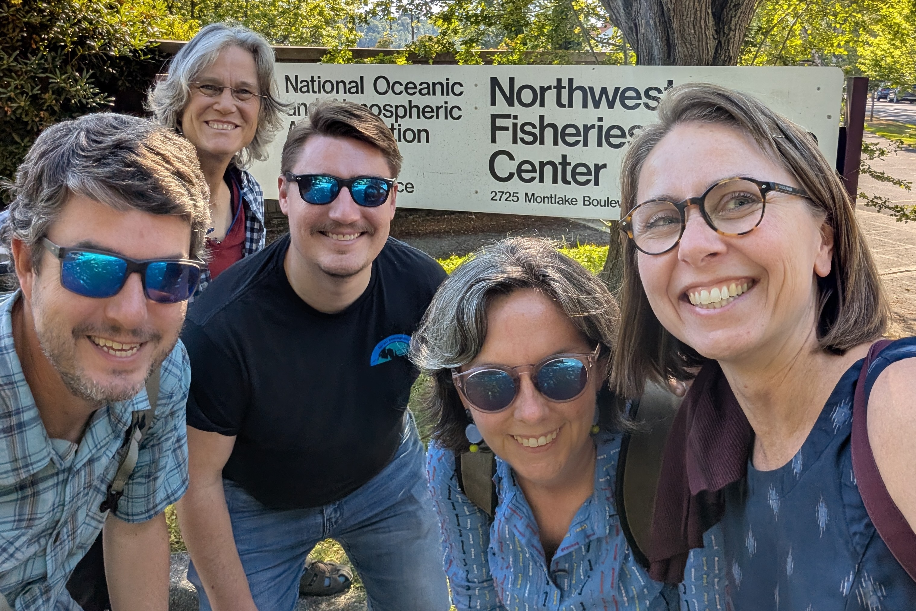 A selfie of 5 smiling people in front of a Northwest Fisheries Science Center sign with a huge deciduous tree in the background.
